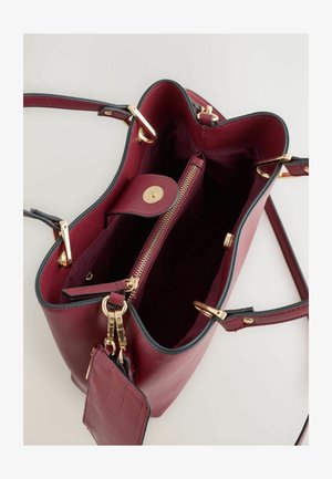 Burgundy nahkalaukku kullanvärisillä yksityiskohdilla, vetoketjullinen osasto ja sisätasku. Varustettu teksturoidulla pinnalla ja kontrastivärillä mustalla reunuksella.
