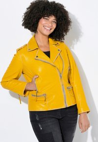Chaqueta de motocicleta de cuero amarillo con cremalleras y detalles plateados, con un cierre asimétrico y dos bolsillos frontales.