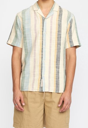 Mann trägt ein kurzärmeliges Hemd mit vertikalen Pastellstreifen und beigen Shorts, steht vor einem einfarbigen Hintergrund.