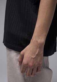 Μαύρο ριμπ πλεκτό πουλόβερ με τελείωμα στη βάση, υφασματένια υφή και ετικέτα λογότυπου στη μία πλευρά. Χέρια τοποθετημένα στο κάτω μέρος.