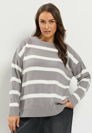 Femme aux longs cheveux bruns portant un pull rayé gris et blanc et un pantalon foncé, posant avec une main dans la poche sur un fond uni.