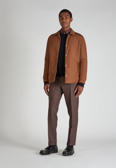 Braune Zip-Jacke, marineblauer Pullover, karierten braune Hose und schwarze Slipper. Glattes Material, Knopfakzente und taillierte Passform.