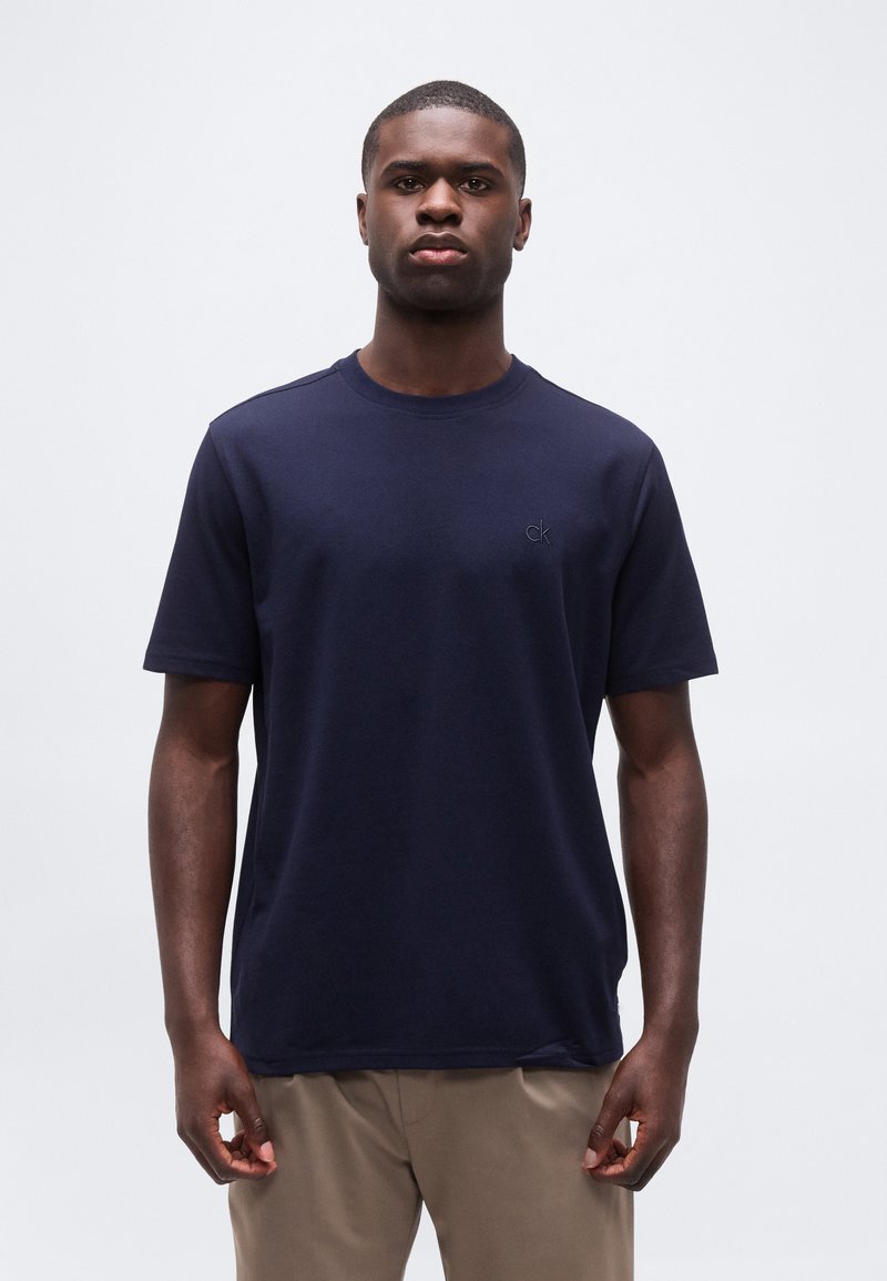 Άνδρας στέκεται προς τα εμπρός φορώντας μπλε ναυτικό μπλουζάκι με μικρό λογότυπο "ck" στο στήθος και μπεζ παντελόνι, με φόντο ένα απλό τοπίο.