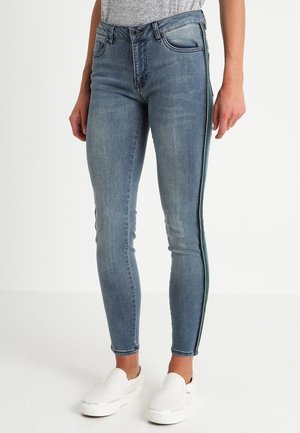 Slim fit blå jeans med mørke sidelister, båret med hvide slip-on sko og en grå top, vist fra talje til fødder.