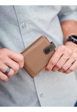 Brown fabric wallet with a smooth texture, featuring a green accent strap and snap button closure, held in a hand against a light shirt.