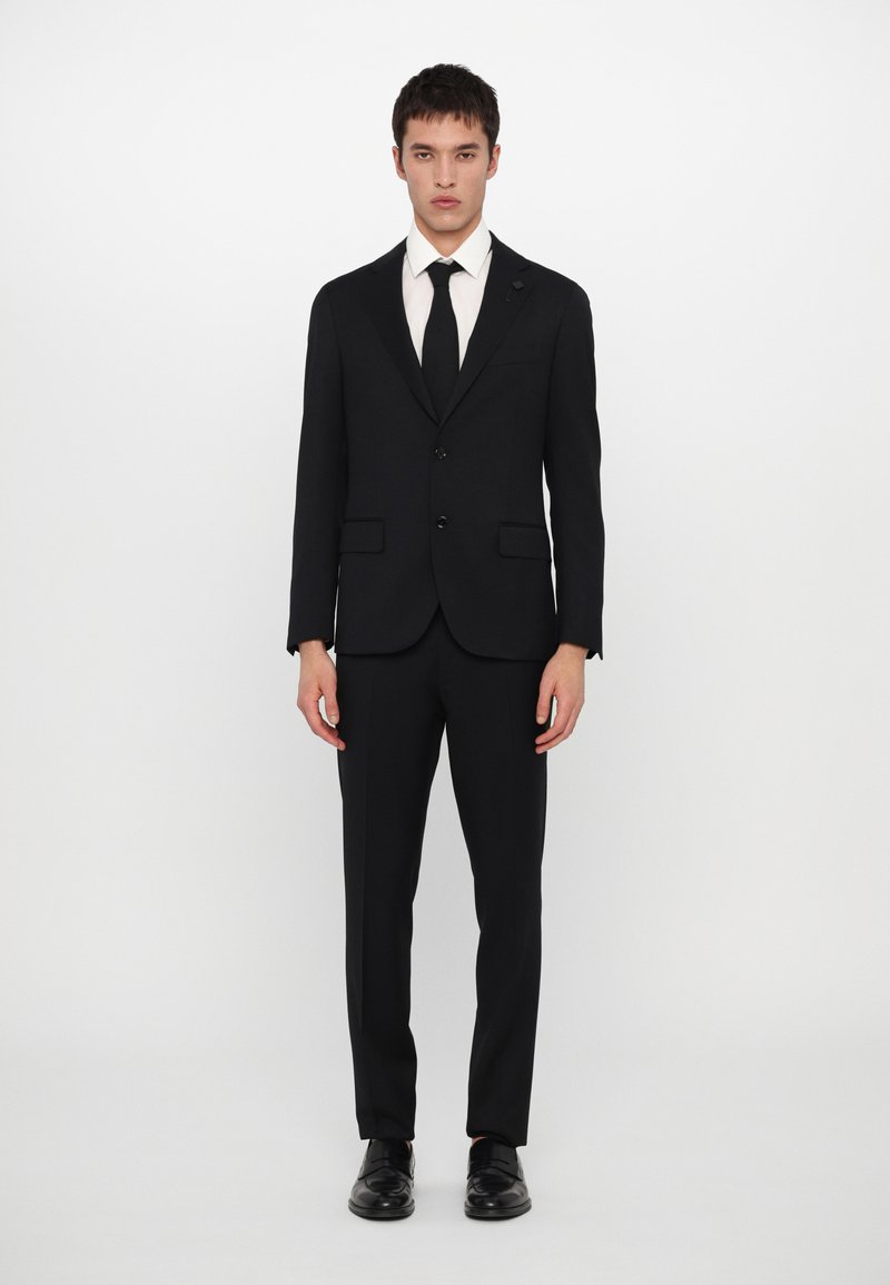 Traje negro con diseño ajustado, que cuenta con dos botones, solapas en pico y bolsillos laterales. Combinado con una camisa blanca y una corbata negra, de acabado satinado.