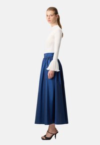 White long-sleeve top with ruffled cuffs paired with a floor-length blue skirt, featuring a smooth texture and elastic waist. Black heeled sandals.