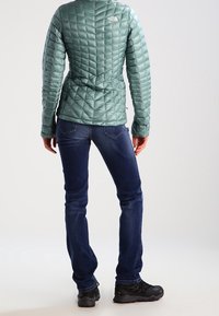 Světlounce zelená prošívaná izolační bunda s přiléhavým střihem, doplněná tmavě modrými džíny s rovnými nohavicemi a černými turistickými botami.