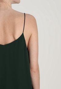 Mörkgrön, ärmlös klänning med V-formad rygg, tunna svarta band och en mjuk, flödande tygstruktur.