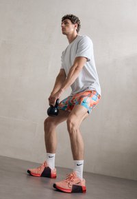 Chaussures de sport roses avec des accents noirs, portées avec des chaussettes blanches et un short coloré arborant un motif abstrait bleu et orange, tenant un kettlebell.