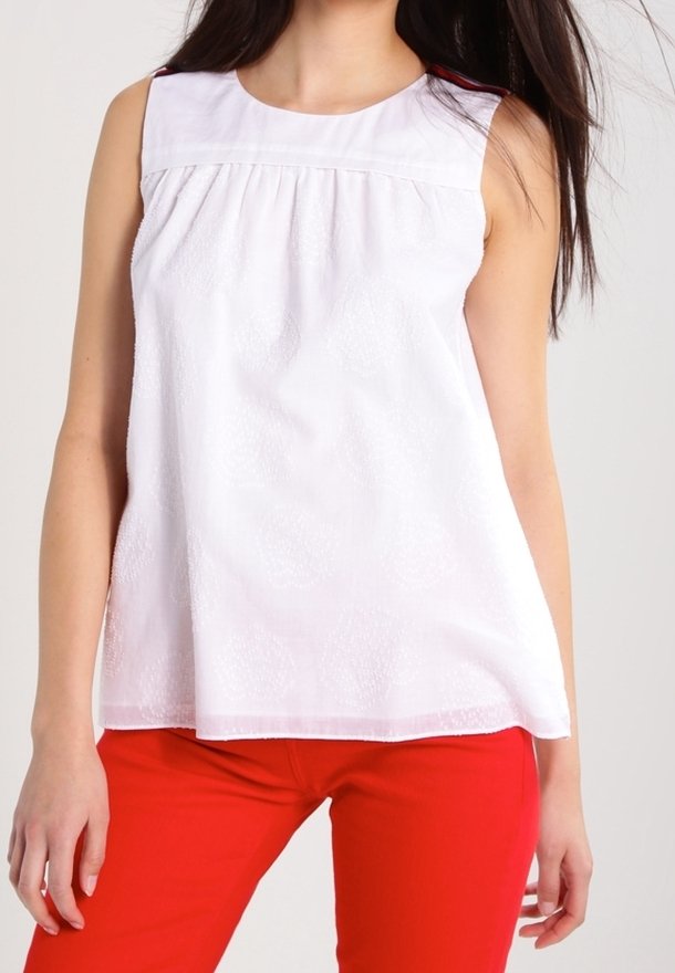 Femme portant un chemisier blanc sans manches avec une texture florale subtile et un pantalon rouge vif, vue du cou aux hanches.