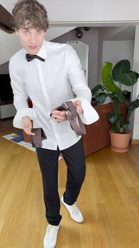 Vit skjorta med breda manschetter, svart fluga, bruna hängslen och smala svarta byxor, kombinerat med vita sneakers. Inomhusmiljö med växter.