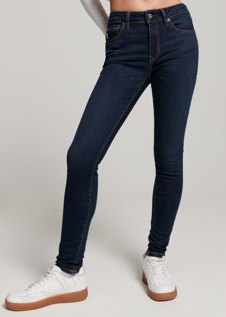 Jeans skinny bleu foncé en denim à la texture lisse, avec des coutures orange et un design classique à cinq poches, associés à des baskets blanches.