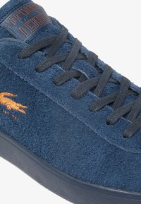 Zapatilla de gamuza azul con superficie texturizada, cordones oscuros y un logotipo naranja en el lateral. Cuenta con puntera redondeada y suela gruesa de goma.