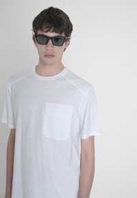 T-shirt bianco a maniche corte con finitura testurizzata e taschino sul petto. Il modello indossa occhiali da sole rettangolari neri su uno sfondo chiaro.