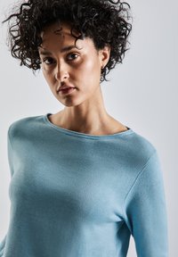 Helles blaues langärmliges Strickoberteil mit rundem Halsausschnitt, das eine glatte Textur und einen figurbetonten Schnitt aufweist. Einfacher, minimalistischer Stil.