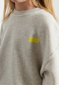Giovane che indossa un maglione grigio chiaro con un piccolo logo giallo "AMERICAN VINTAGE" sul petto, viso parzialmente visibile e capelli biondi.