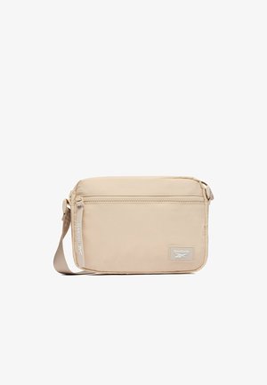 Bolso bandolera rectangular beige con bolsillo frontal con cremallera, correa de tela ajustable y parche con el logo de Reebok en la esquina inferior derecha.