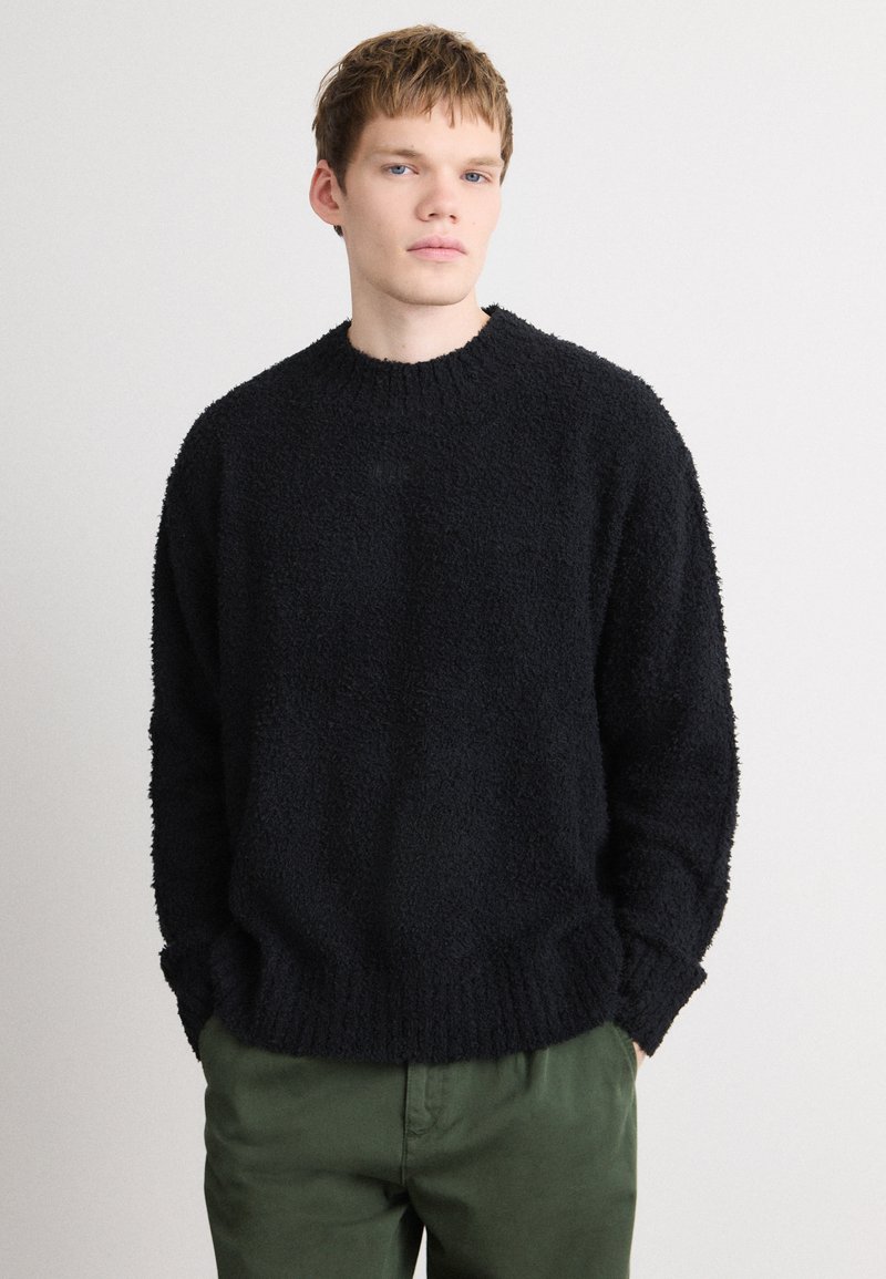 Pulover negru, tricotat cu textură, cu guler rotund și manșete cu nervuri, asociat cu pantaloni de culoare verde măslin. Material moale și pufoasă, cu o croială lejeră.