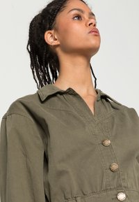 Vestido camiseiro de algodão verde-azeitona com colarinho, cinco Botões e cintura com cinto. Apresenta um tecido texturizado e silhueta estruturada.