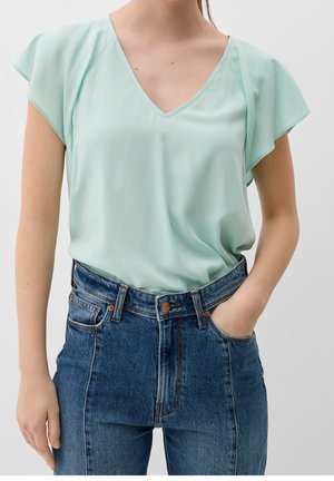 Femme portant un chemisier vert clair à col en V avec des manches volantées, rentré dans un jean bleu taille haute, une main dans la poche.