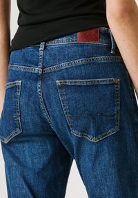 Skats no muguras uz personu, kas valkā zilas džinsu bikses ar redzamām aizmugurējām kabatām un brūnu zīmola etiķeti, kopā ar melnu augšdaļu.