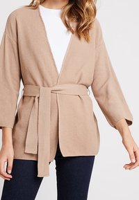 Beige stickad cardigan med bälte, V-ringning, vida ärmar och rak passform. Mjuk textur, minimalistisk design, idealisk för lager-på-lager.