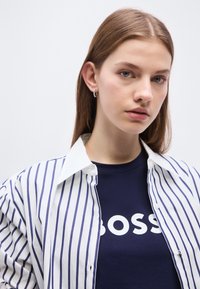 Νεαρή γυναίκα με ίσια καστανά μαλλιά φοράει ασημένια σκουλαρίκια κρίκους, μπλε πουκάμισο με το κείμενο «BOSS» και ανοιχτό λευκό πουκάμισο με μπλε κάθετες ρίγες.