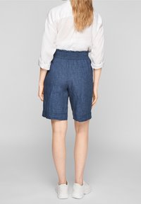 Pantalones cortos de lino azul con cintura fruncida, ajuste tapered y puños enrollados. Combinados con una camisa blanca de manga larga y zapatillas blancas.