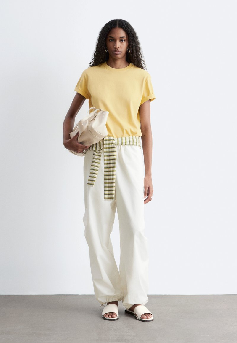 Femme aux cheveux bouclés portant un t-shirt jaune, un pantalon blanc avec un pull rayé noué à la taille, tenant un sac beige, debout sur un sol gris.