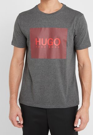Mężczyzna nosi szarą koszulkę z krótkim rękawem z dużym czerwonym prostokątnym logo z napisem "HUGO HUGO BOSS" na piersi.
