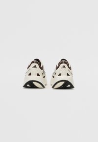 Baskets blanches et marron avec un dessus texturé, présentant des accents noirs et un design de semelle sculptée. Logo Adidas sur le talon.