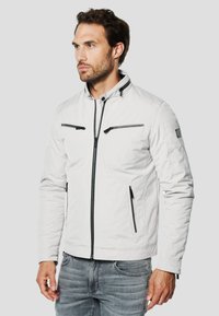 Witte jack met ritssluiting en zwarte ritssluitingaccenten, voorzien van zijzakken en eenstaande kraag. Gemaakt van een glad, lichtgewicht materiaal.