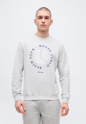 Young man wearing light gray sweatshirt and matching sweatpants, standing against plain white background.