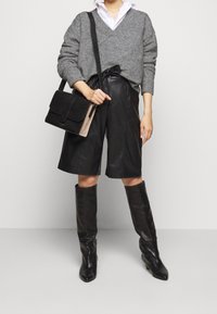 Maglione grigio oversize, pantaloni corti neri in pelle, stivali neri sopra il ginocchio e una borsa bicolore con un accento testurizzato. Sfondo neutro.