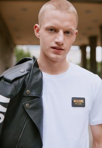 Veste en cuir noir de motard avec quincaillerie argentée, drapée sur un t-shirt blanc avec un patch rectangulaire Moschino Milano.