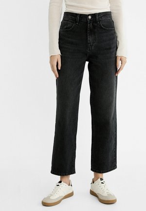 Person iført sort jeans med højtaljede og lige ben, hvid langærmet top og hvide sneakers med lysebrune såler på en ensfarvet baggrund.