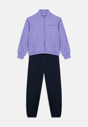 Lilla jakke med glidelås og krage, sammen med svarte bukser. Jakken har raglanermer, elastiske mansjetter og en liten logo.