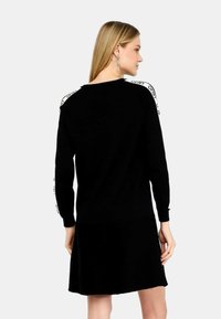 Abito nero con maniche lunghe, caratterizzato da dettagli testuali bianchi lungo le spalle. Morbido tessuto a maglia, forma ad A e design fino al ginocchio.