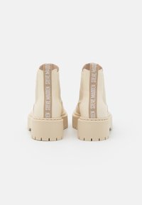 Bottines beiges avec une finition lisse, semelle épaisse et un texte proéminent sur le talon. Panneaux élastiques sur le côté pour un enfiler facile.