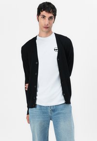 Cardigã preto com botões, usado sobre uma t-shirt branca com um pequeno gráfico. Modelo posa contra um fundo branco simples.