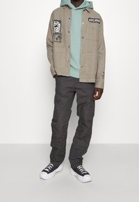 Beige Button-up-Jacke mit Taschen, darunter ein grüner Hoodie, graue Cargo-Hosen und schwarze Sneaker mit weißen Sohlen.