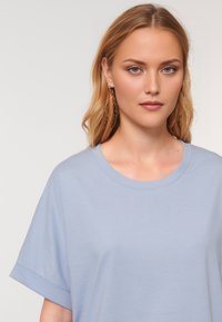 Portrait of a young woman with long, wavy hair, wearing a light blue t-shirt and subtle earrings, posing against a plain background.