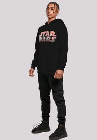 Felpa nera con cappuccio che presenta una grafica multicolore di "Star Wars" sul petto, abbinata a pantaloni cargo neri e sneakers alte nere.