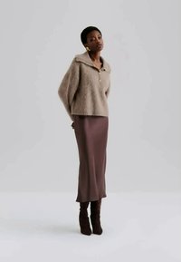 Beige, fuzzy sweater med brede ærmer og krave, kombineret med et langt, brunt satin-nederdel og knæhøje brune støvler. Modellen står med hænderne på hofterne.