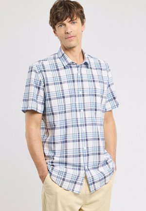 Homme portant une chemise à manches courtes blanche et bleue à carreaux avec un pantalon beige, debout les mains dans les poches sur un fond uni.