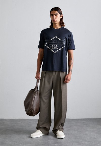 Marineblauw katoenen t-shirt met een grafisch ontwerp met "GA," gecombineerd met beige wijde broek en een bruine leren tote bag. Witte sneakers.