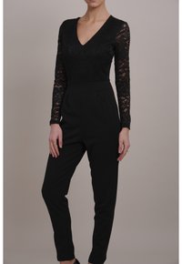 Czarny kombinezon z koronkową bluzką z długim rękawem i dekoltem w kształcie litery V oraz gładkimi, zwę�żanymi nogawkami, tworzącymi dopasowaną sylwetkę.