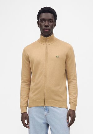 Mann trägt einen beigen zip-up Pullover mit kleinem grünem Logo auf der Brust und hellblauen Jeans, steht vor einem einfarbigen weißen Hintergrund.