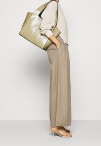 Grand sac fourre-tout vert olive avec texture crocodile, garniture noire, porté avec un pull beige clair et un pantalon ample beige.